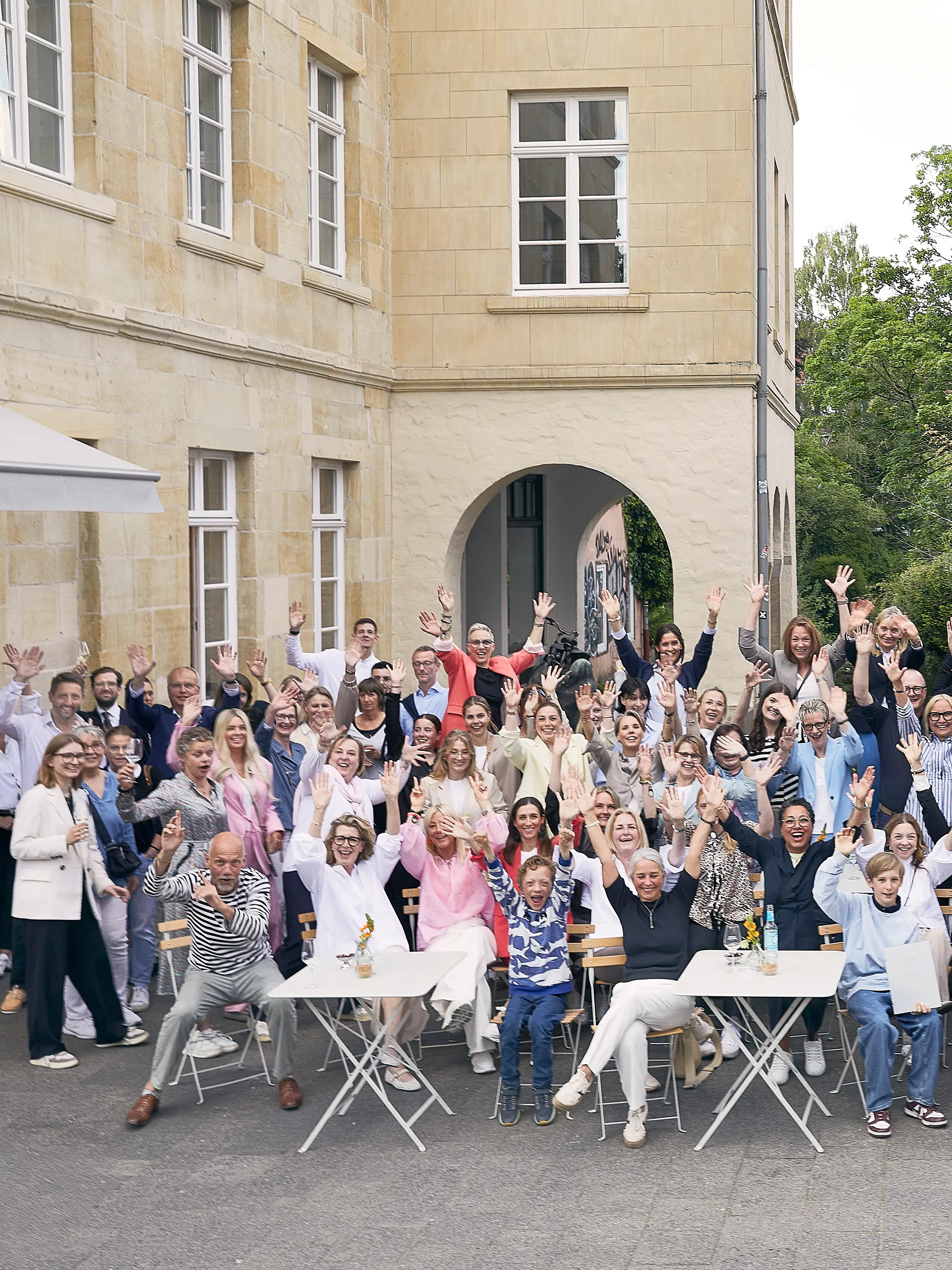 Mitarbeiterinnen und Mitarbeiter sitzen und stehen vor einem hellen Gebäude und winken in die Kamera.