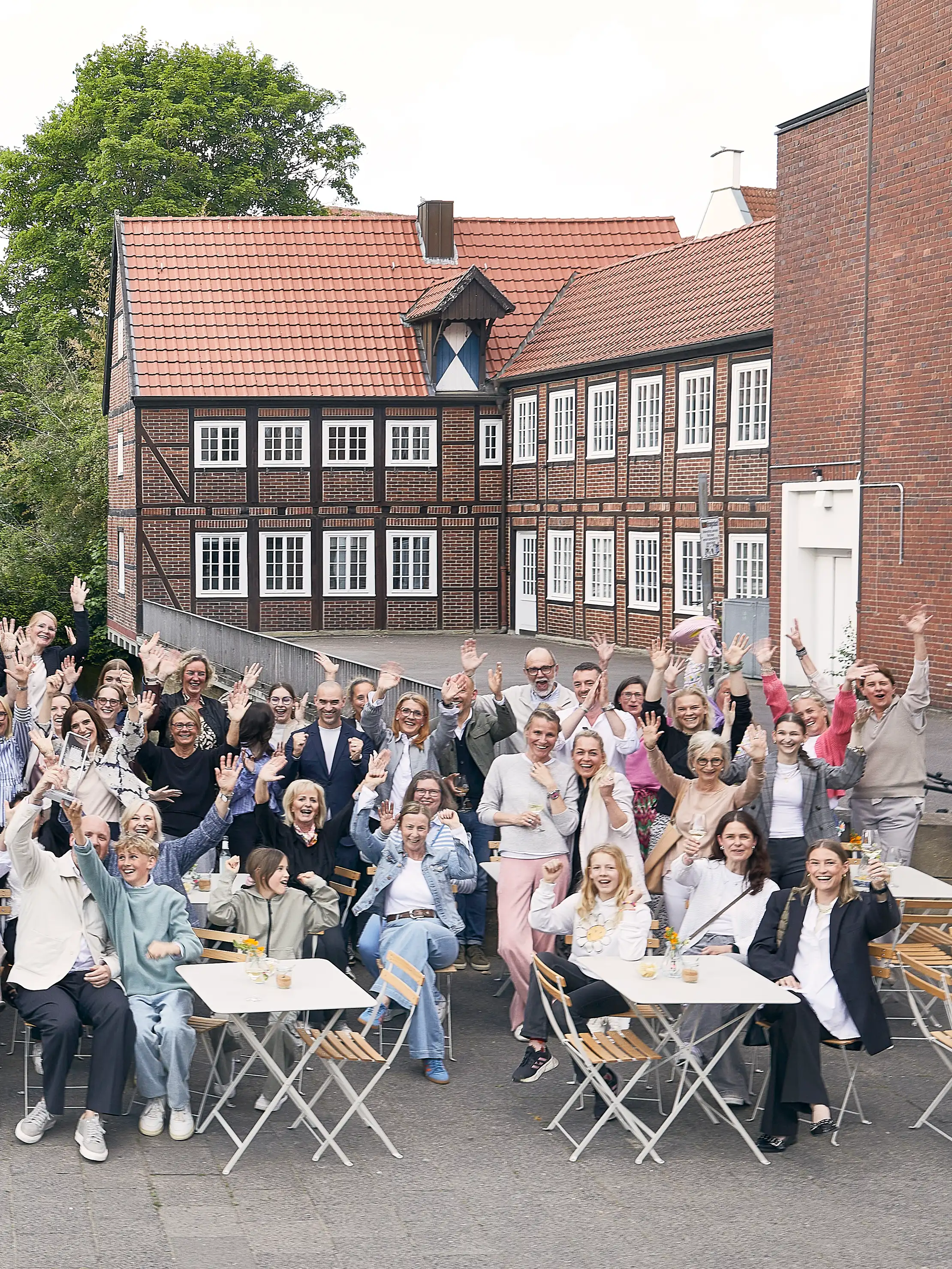 Mitarbeiterinnen und Mitarbeiter sitzen und stehen vor einem hellen Gebäude und winken in die Kamera.